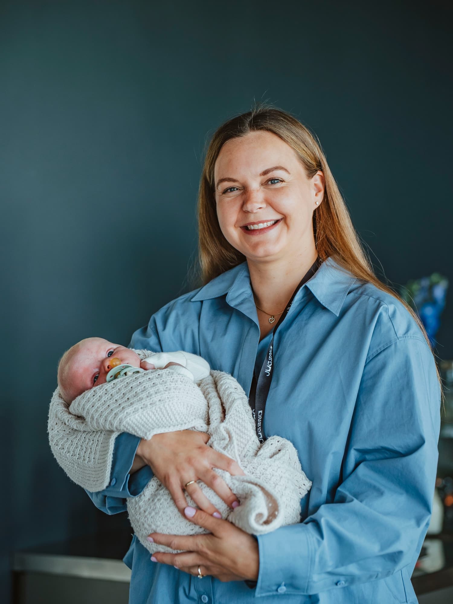 Andrea Dyb smiling, holding a baby wrapped in a knitted blanket, in a blue shirt against a dark wall